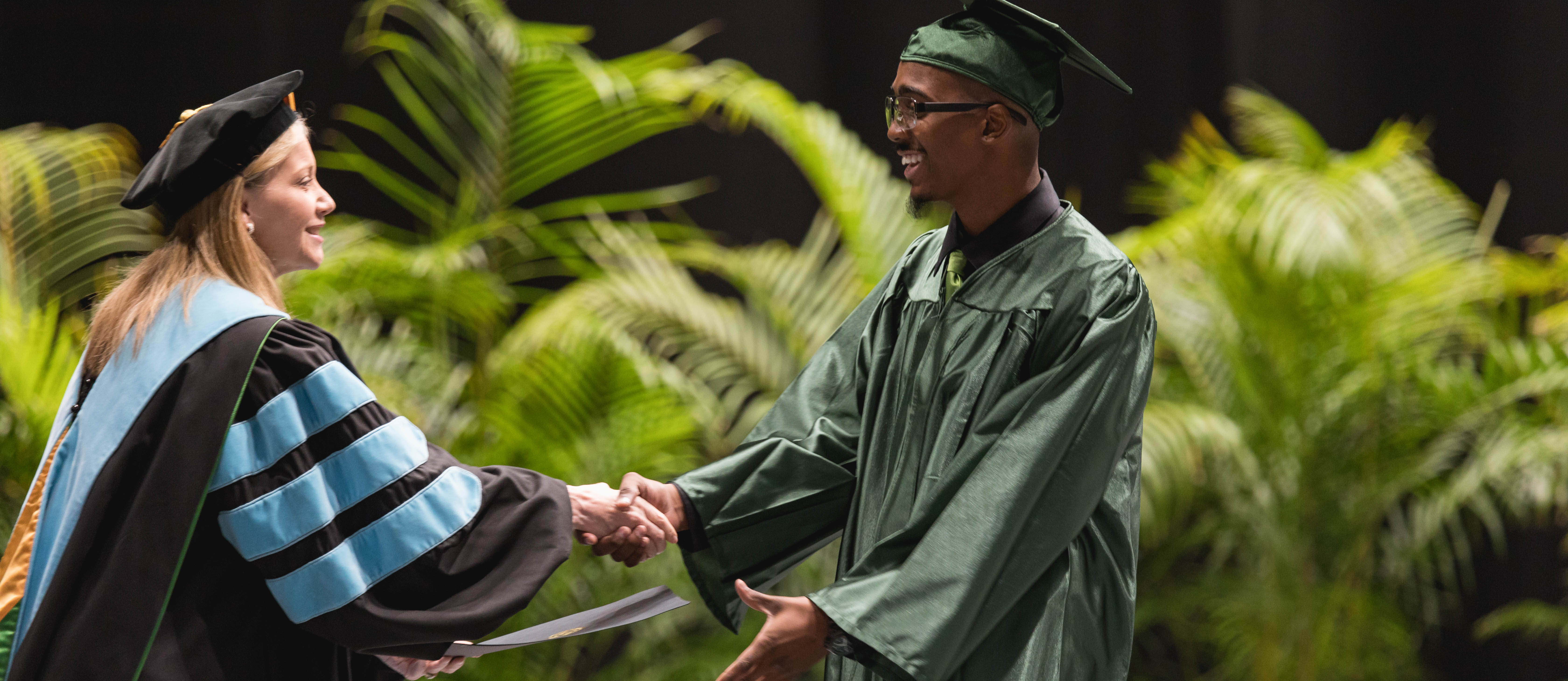 adult high school student in cap and gown shaking hands with President Dr. Deitemeyer as they walk across the stage and accept their diploma