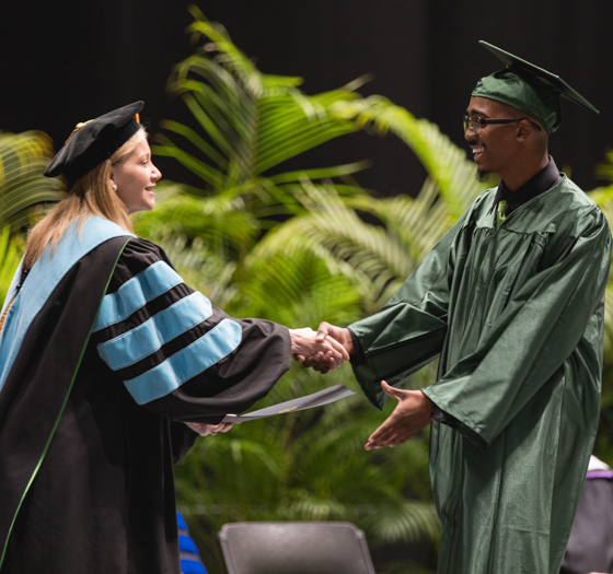 adult high school student in cap and gown shaking hands with President Dr. Deitemeyer as they walk across the stage and accept their diploma