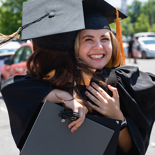 two graduates smiling and hugging in their caps and gowns in a parking lot