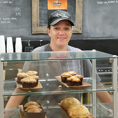 student smiling behind the counter in front of muffins at Aspire Bakery and Bistro