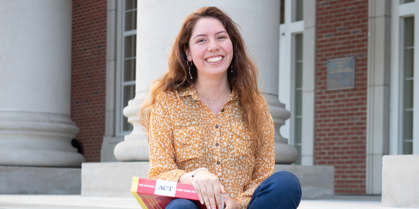 student with long hair and holding ACT prep book smiling on outdoor stairs