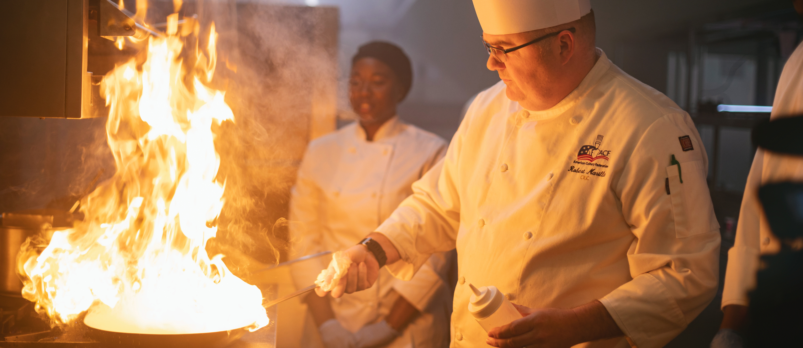 chef shaking a skillet with a large flame as his student watches