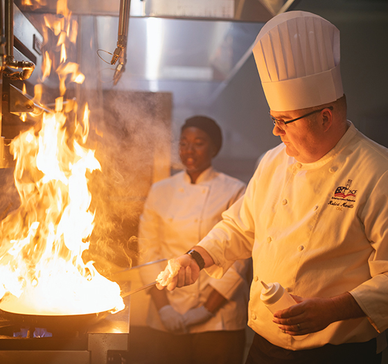 chef shaking a skillet with a large flame as his student watches