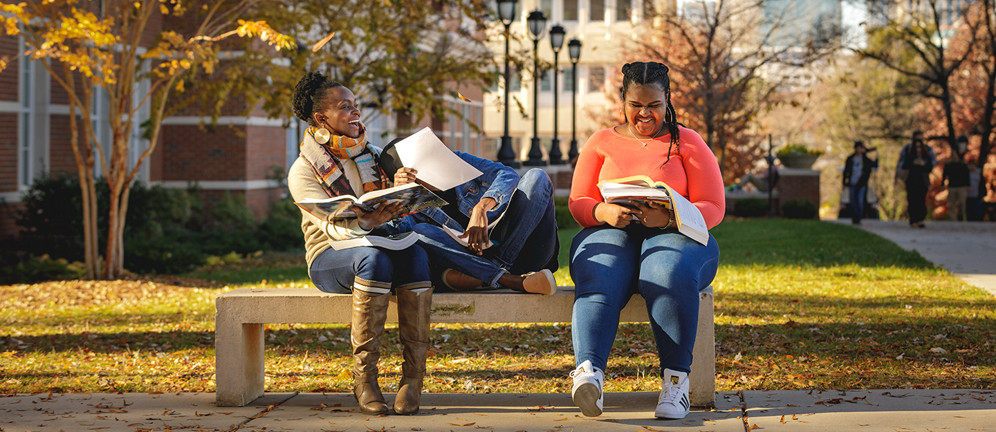 three female students smiling and laughing sitting on a bench as they study on Central Campus with the Charlotte skyline behind them