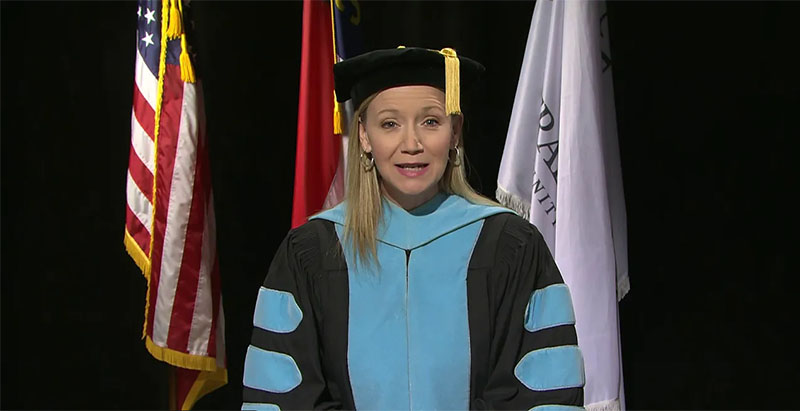 Central Piedmont President Dr. Deitemeyer addressing graduates wearing graduation regalia with American, NC, and Central Piedmont flags behind her