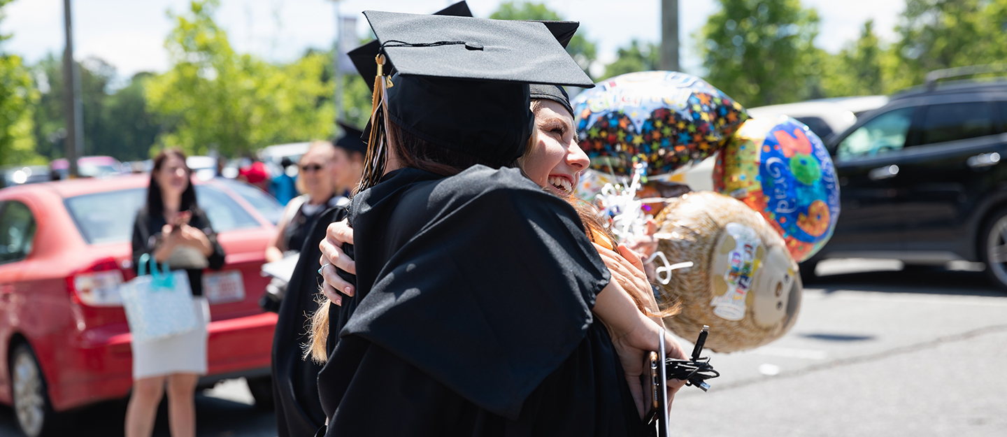 two graduates smiling and hugging in their caps and gowns in a parking lot