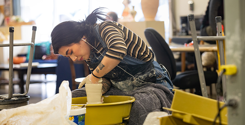 a student in overalls working at a pottery wheel