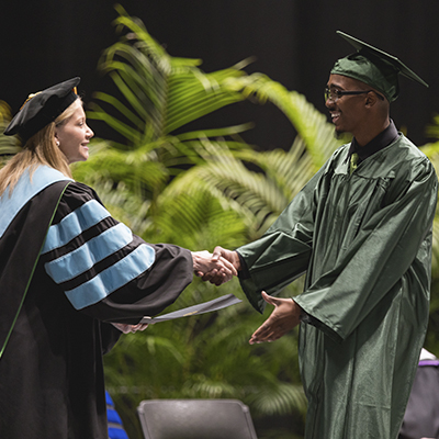 adult high school student in cap and gown shaking hands with President Dr. Deitemeyer as they walk across the stage and accept their diploma