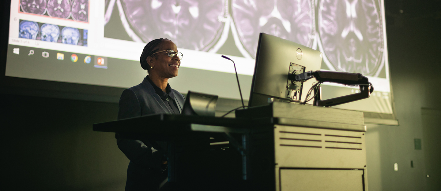 instructor at podium in front of screen showing slides of the human brain