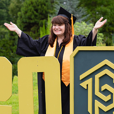 student in cap and gown raising hands excitedly behind class of 2020 sign at drive-through graduation