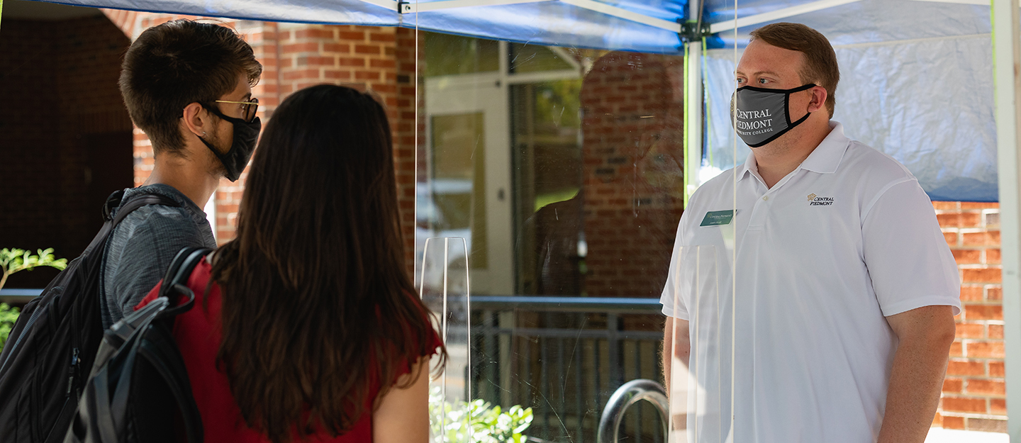 a Central Piedmont Student Life staff member wearing a Central Piedmont mask behind a Plexi-glass shield helping students wearing masks on campus