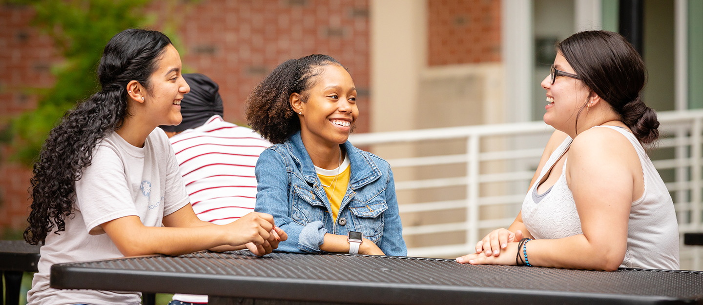 three students sitting outside at a picnic table smiling and talking