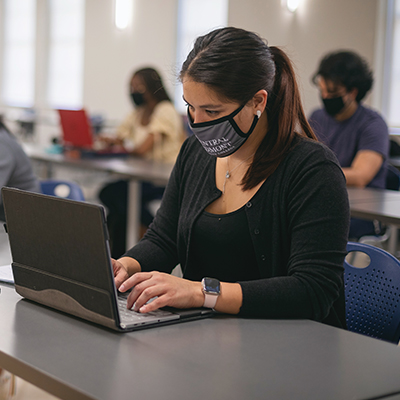 student ambassador sitting in front of laptop in classroom wearing a mask