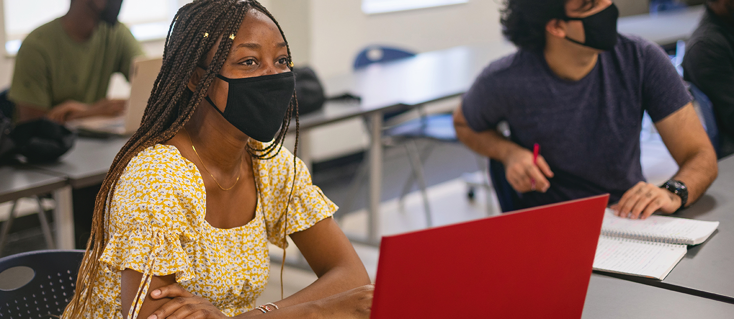 student wearing mask with laptop on desk listening to instructor