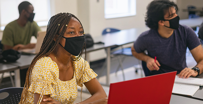 student wearing mask with laptop on desk listening to instructor