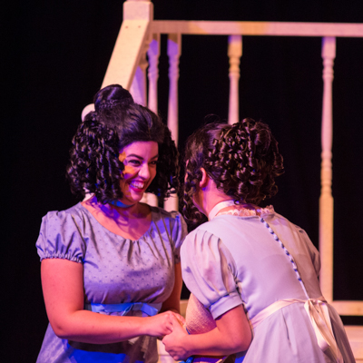 two actors in 18th century dresses giddly holding hands and talking