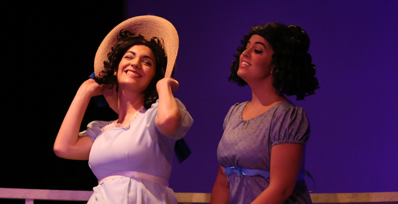 two actors in 18th century dresses smiling and talking, one wearing a floppy hat