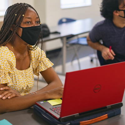 student wearing mask with laptop on desk listening to instructor