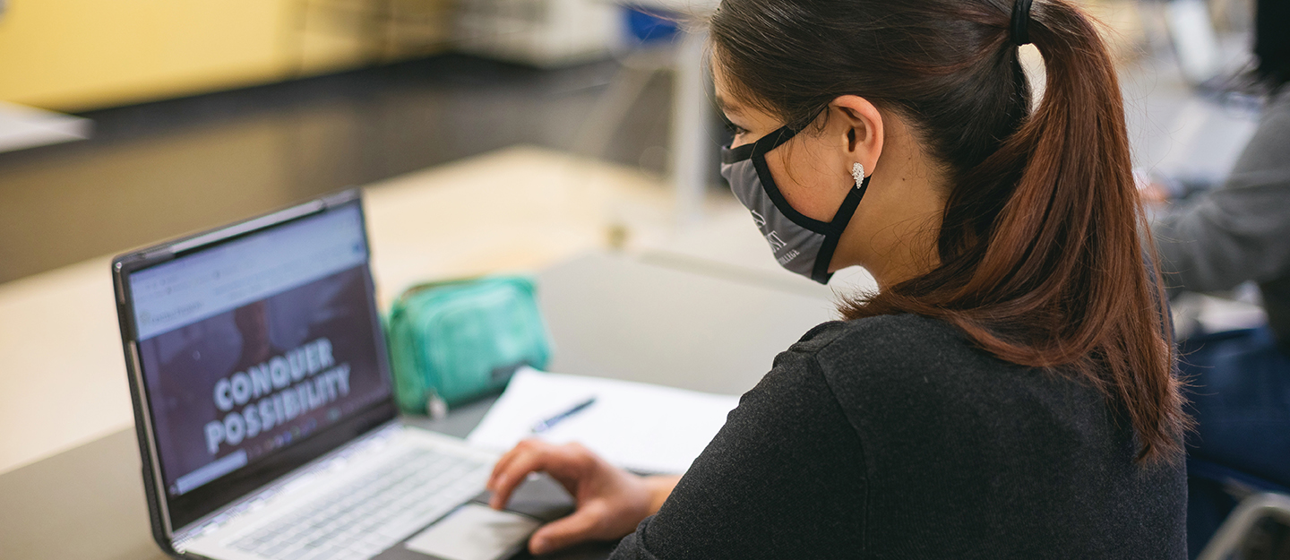 student with ponytail wearing mask on laptop in a classroom