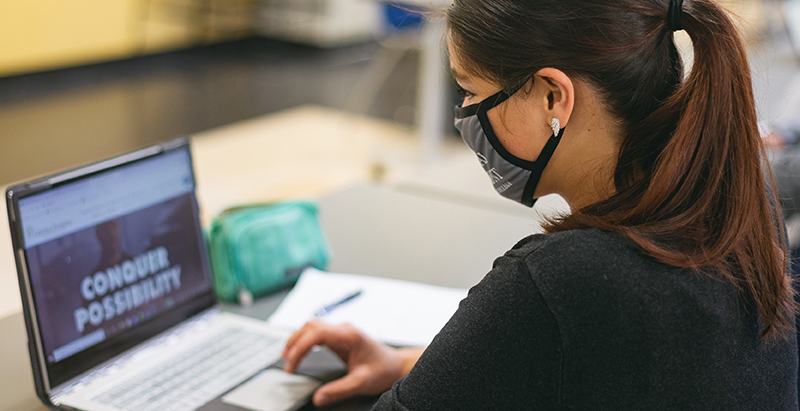 student with ponytail wearing mask on laptop in a classroom