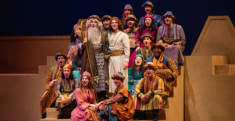 chorus members from joseph and the amazing technicolor dreamcoat sitting on steps smiling and singing