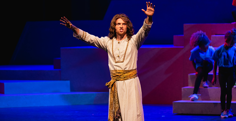 Joseph and the amazing technicolor dreamcoat actor on stage in biblical dress with arms raised, singing