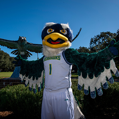 UNC Wilmington mascot Sammy Seahawk with wings spread out standing on campus in front of a hawk statue