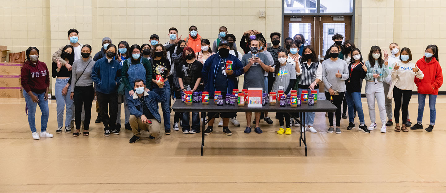 large group of diverse opportunity scholars standing by table of peanut butter and jelly jars at service event in Worrell gym