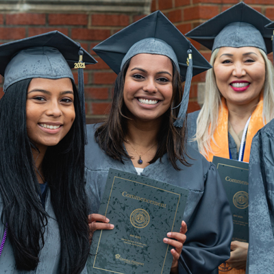 three smiling Central Piedmont graduates in their caps and gowns holding up their commencement programs