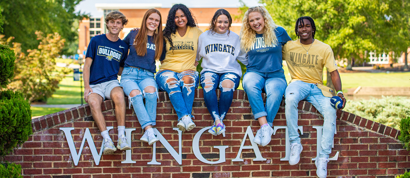 Wingate University and Central Piedmont diverse group of students sitting on Wingate University sign