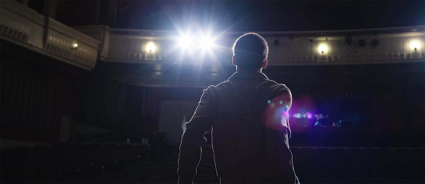 Backlit photo of a Central Piedmont student standing on a stage looking out at the audience