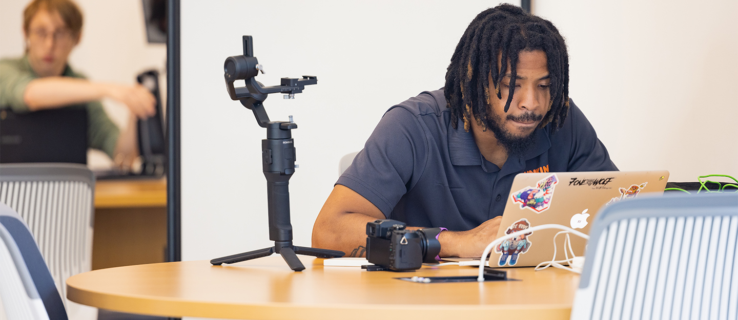 A male student working on his laptop with a camera nearby