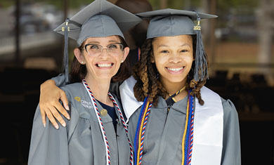 Two smiling graduates, one with an arm around the other, wearing Central Piedmont cap and gown and various honors cords.