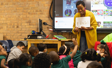 Smiling teacher at the front of a classroom looks at student with a hand raised