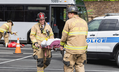 Two firefighters carry a stretcher between them with a police car in the background.