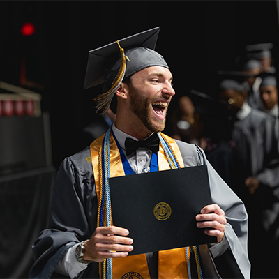 One person in cap and gown on stage holding their diploma 