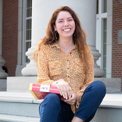 student siting on steps with test prep book smiling into camera