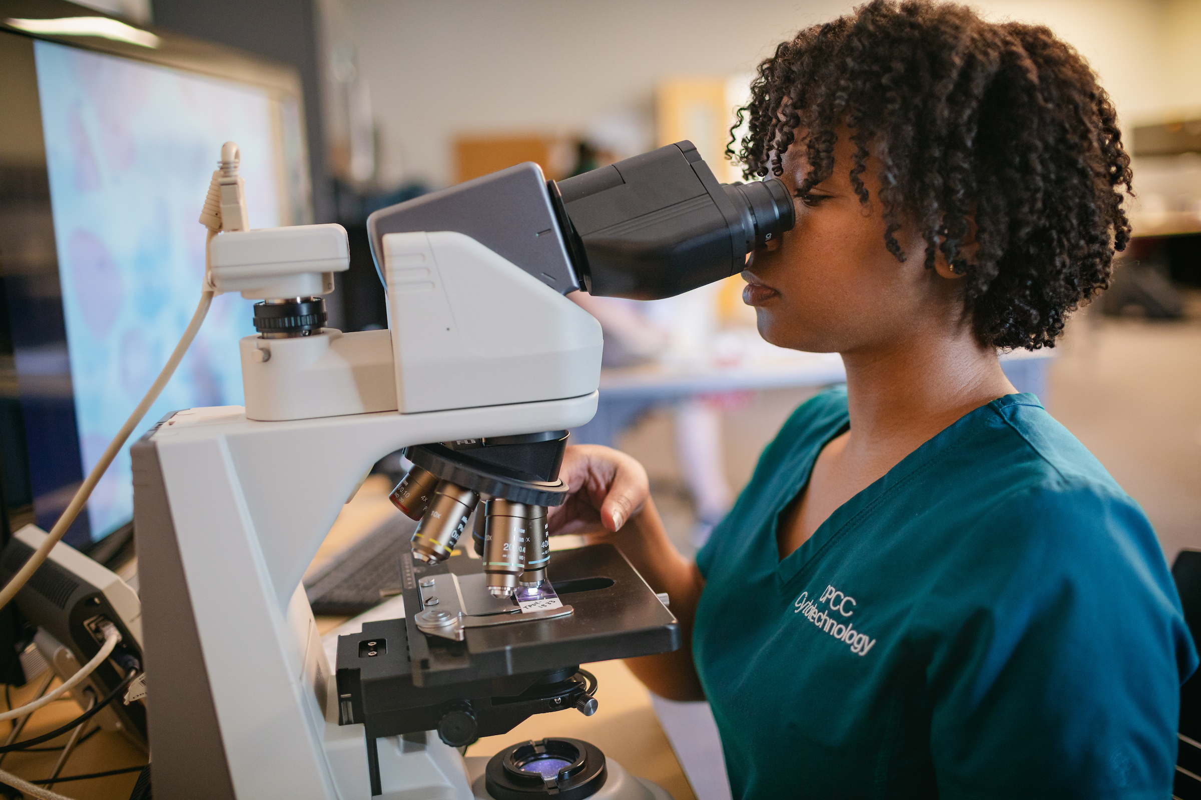 Central Piedmont student wearing cytotechnology scrubs looking through microscope in lab.