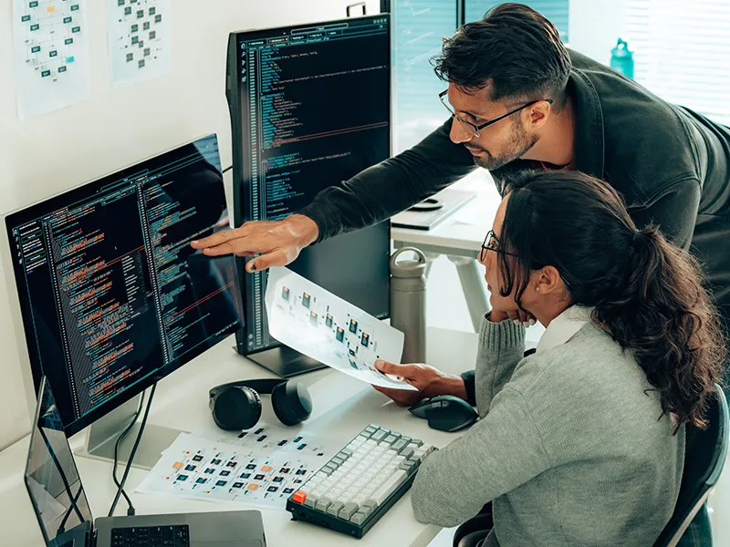 Two IT staff reviewing code together at a multi-monitor workstation