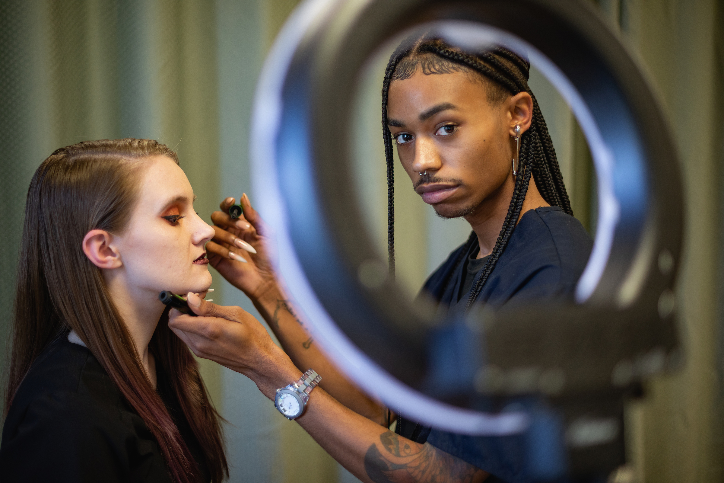 Central Piedmont student looking through ring light into camera while about to apply mascara to fellow student.