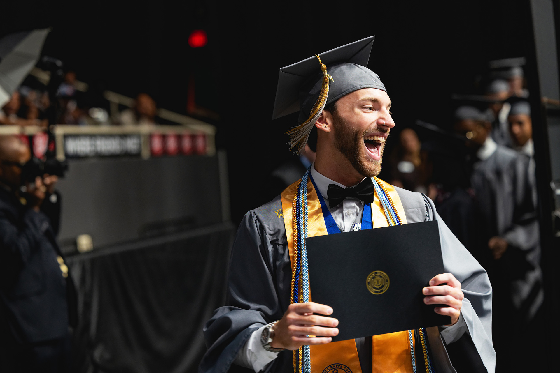 Smiling Central Piedmont wearing a black graduation cap and gown and honors cords holding diploma at graduation.