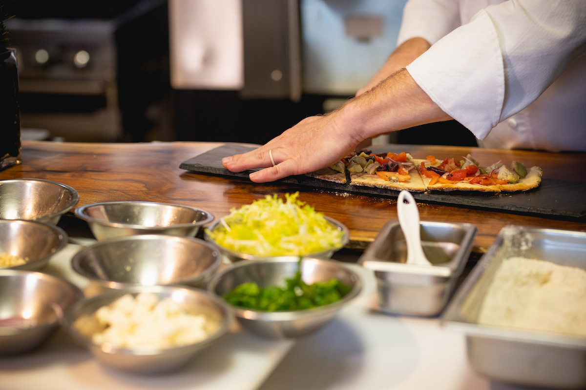 Closeup of chef’s hands cutting a flatbread with small bowls of ingredients in foreground.