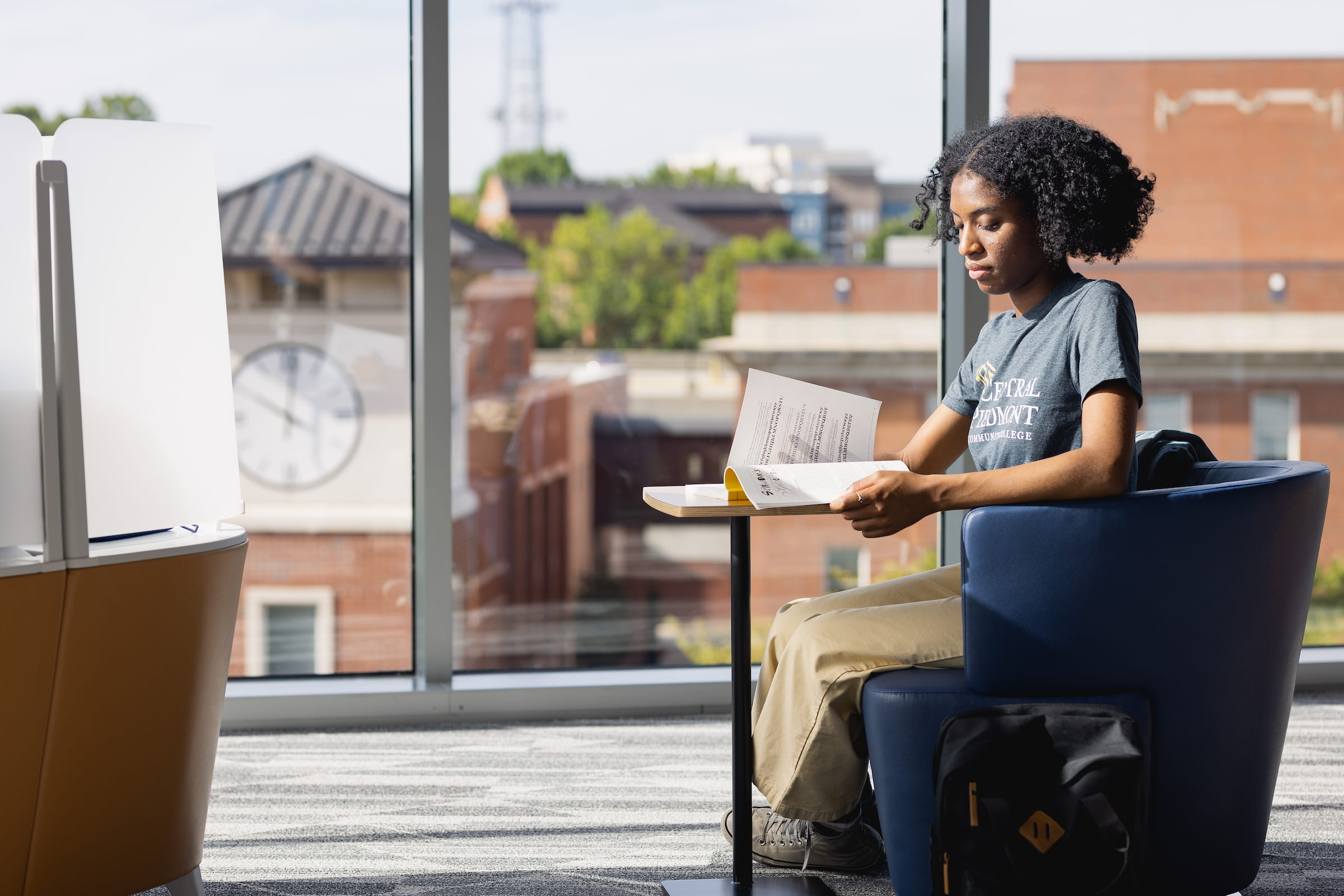 Central Piedmont student reading book at small table in common area with brick campus buildings visible through windows.