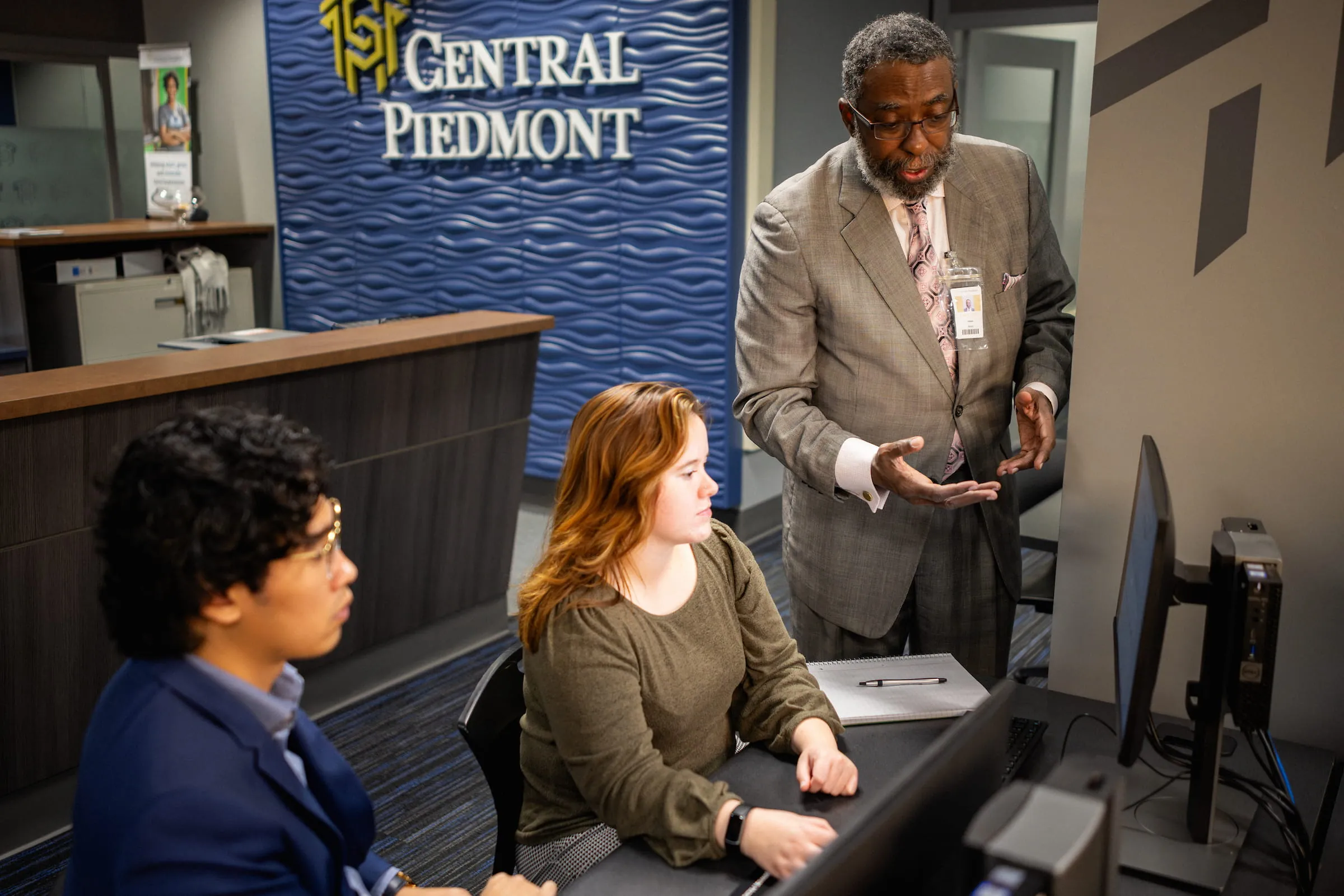 Staff member talking with two colleagues at a desk beneath a Central Piedmont sign