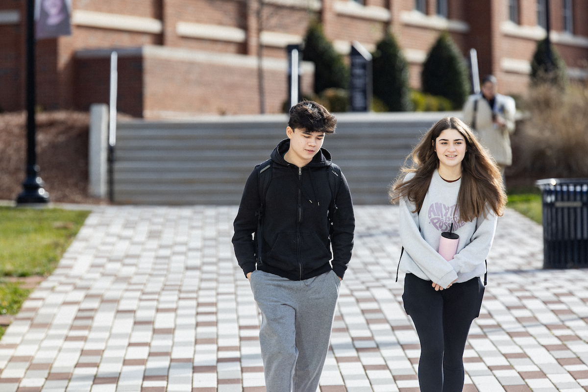 Two Central Piedmont students walking outside on paved walkway with stairs and Central Campus buildings in background.