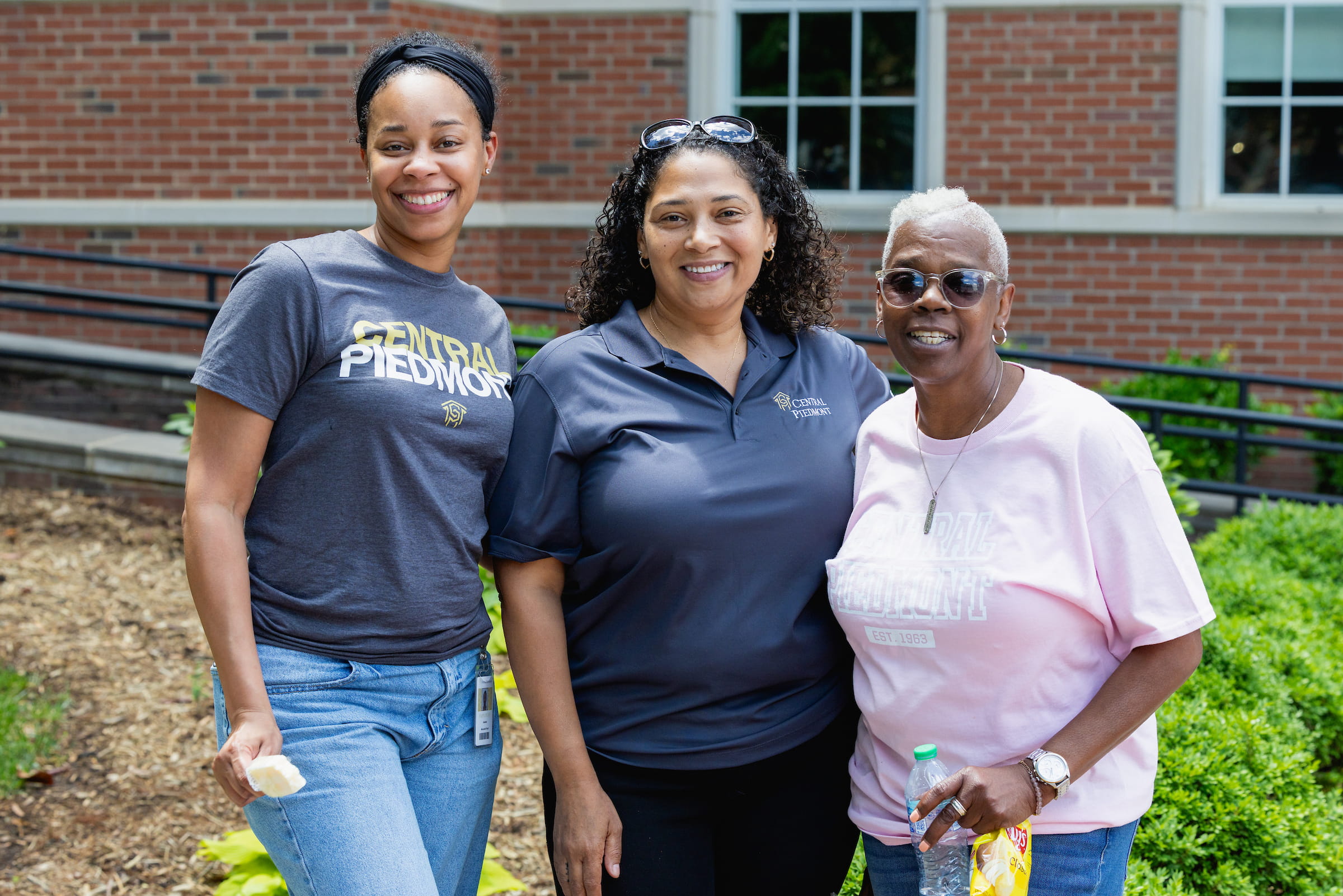 Three Central Piedmont staff members in college shirts smiling together outside a brick building