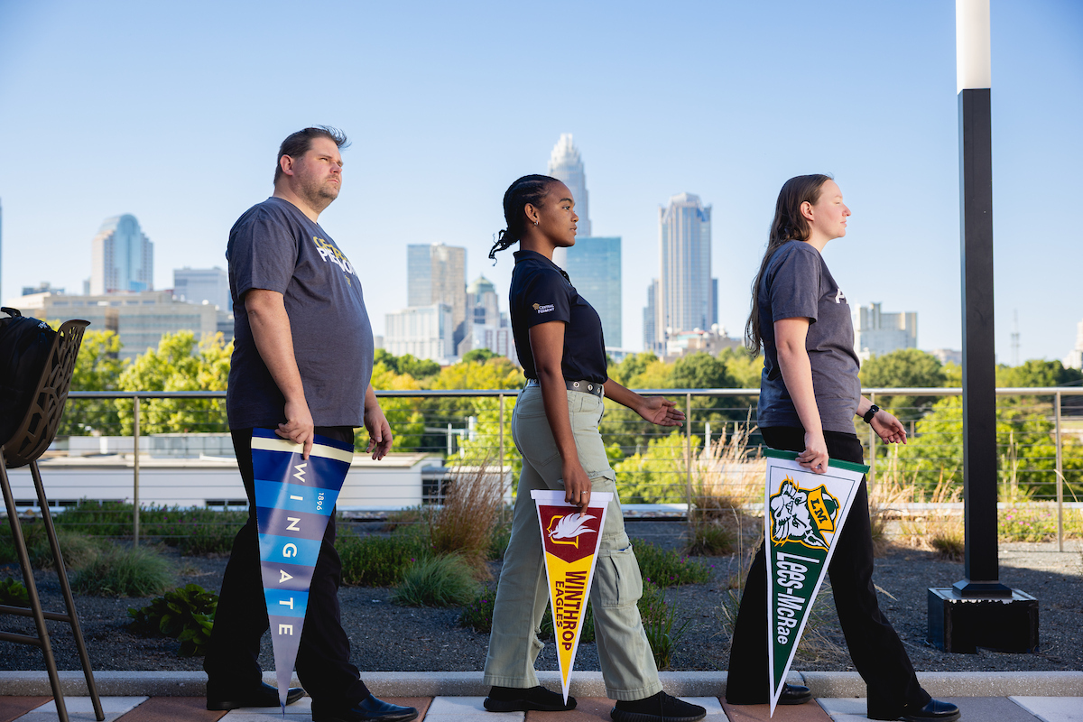 Three students walking in a row holding university pennants against the Charlotte skyline