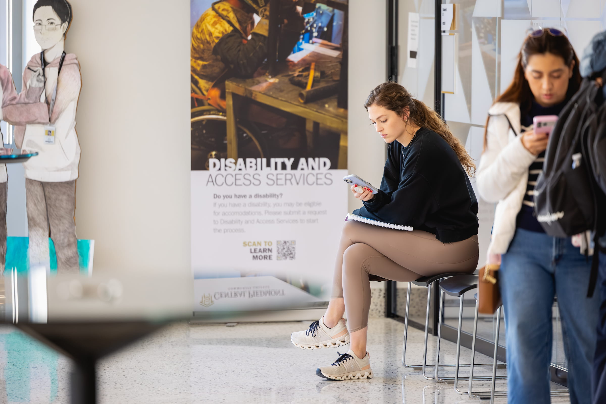 Central Piedmont student on phone and sitting on chair in front of sign that reads “Disability and Access Services.”