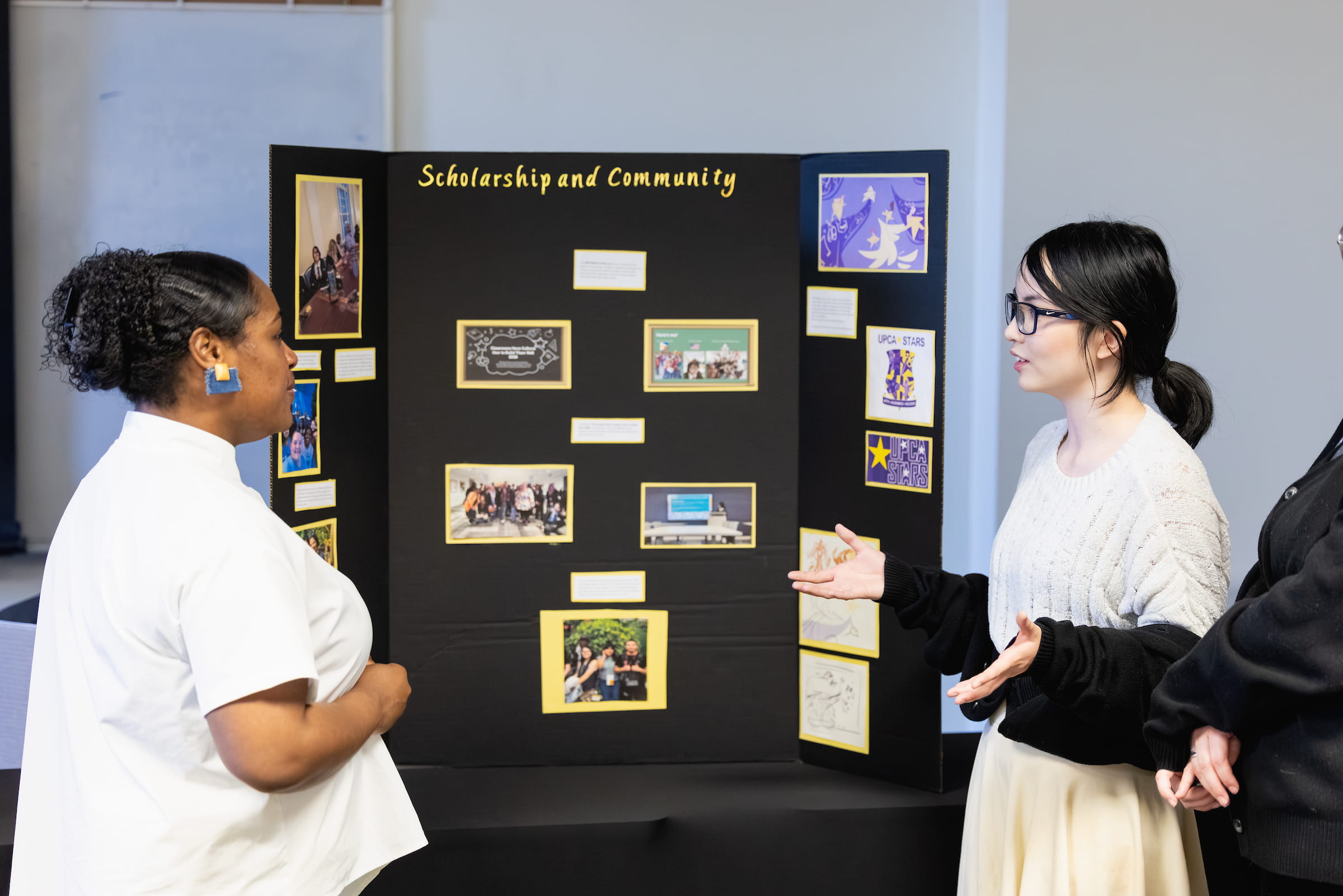 Central Piedmont student presenting trifold board reading “Scholarship and Community” to person listening intently.