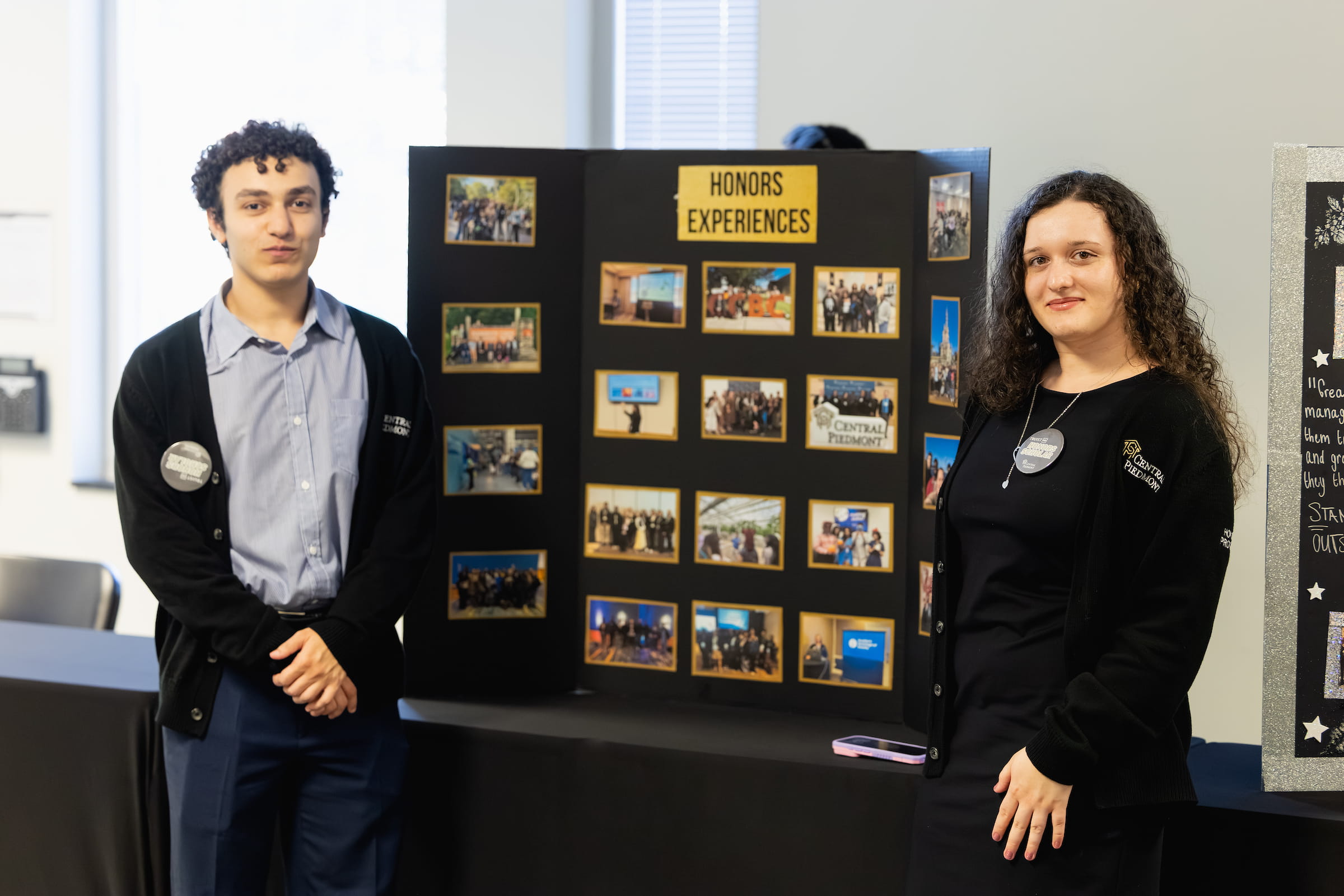 Two students smiling for camera in front of trifold board reading “Honors Experiences” with photos on display.